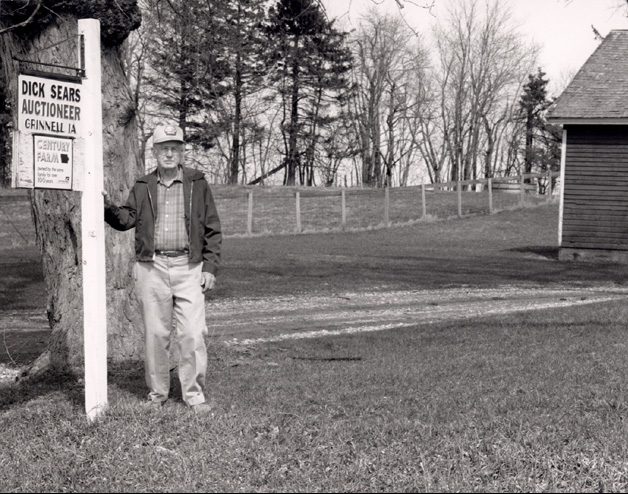 Man standing, outside, by the sign "Dick Sears Auctioneer Grinnell IA"